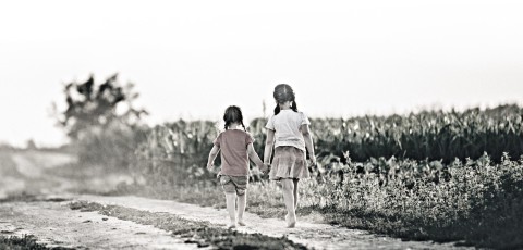 Two sisters walk down a dirt road near a corn field.